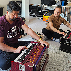 Two men sit on the floor, each with a harmonium, one finger on its keyboard and a hand on the bellows at the rear. The man at left is the student; he is a stocky man with light brown skin, short black hair, beard and mustache. At right is his teacher, a smaller, slender man with Asian features, the ghost of a mustache and thinning dark hair pulled back.