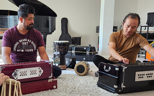 Two men sit on the floor, each with a harmonium. The man on the left, the student, is watching his teacher play the harmonium.