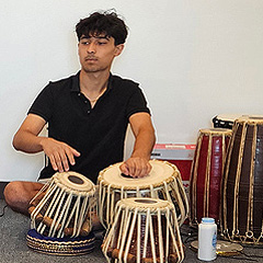 A young man sits cross-legged on the ground, behind two drums which he is playing with his hands.