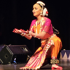 A young woman in a brightly-colored sari kneels on the dance floor, her hands held in front of her in a gesture of yearning. Her face has a desperate, imploring expression.