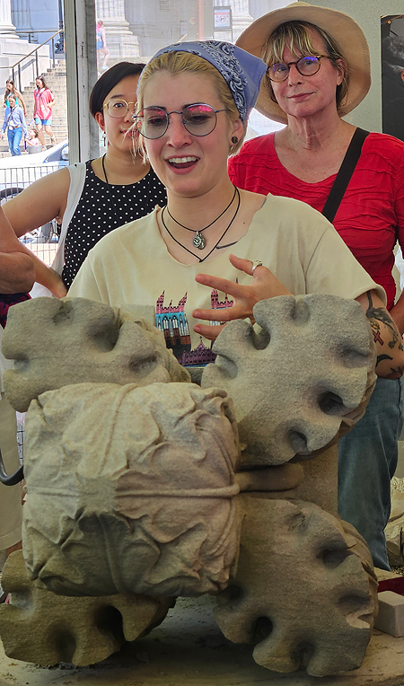 A young woman stands behind a large concrete cabbage, smiling and talking animatedly. She is wearing large round blue-tinted glasses, a yellow National Cathedral T-shirt with raw neckline and sleeves, and large colorful tattoos on her arm.