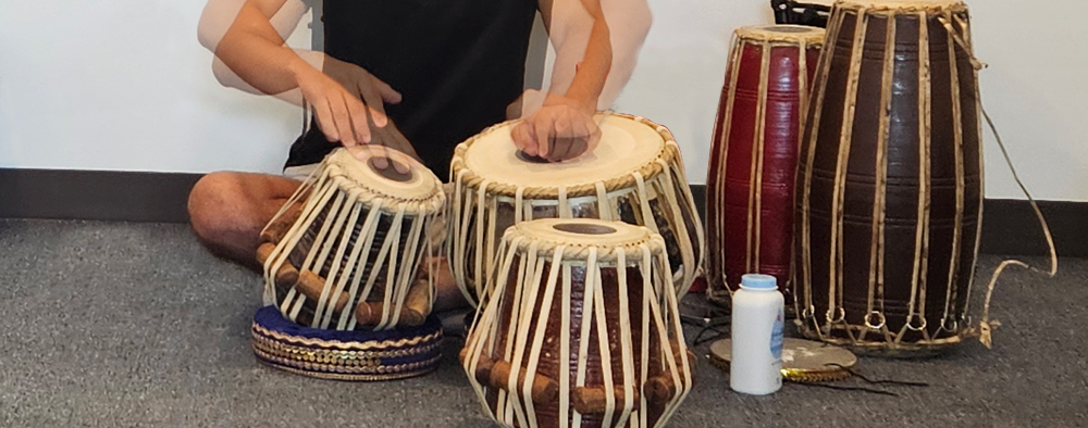 Biplab Khatiwada plays tablas, in a double exposure that creates a sense of motion in Biplab's arms and hands.  He sits cross-legged on the floor behind two drums, each about a foot high, one narrower and higher pitched than the other. Three larger, taller drums are off to the side.
