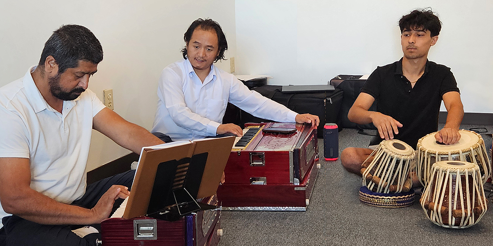 Biplab Khatiwada sits cross-legged on the floor behind two drums, playing them with his hands. Each drum is about a foot high, one narrower and higher pitched than the other. Three larger, taller drums are off to the side.