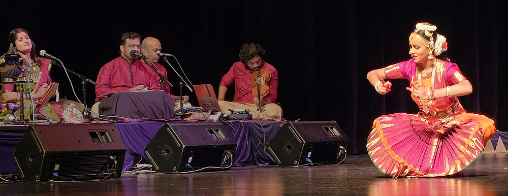 A young woman performs a classical Indian dance.She is crouching with her arms out in front of her as if she holds a box, and she looks worried. At left are four musicians sitting cross-legged on a low platform. The dancer's dress is magenta and orange, with a pleated skirt, and decorations on her head.