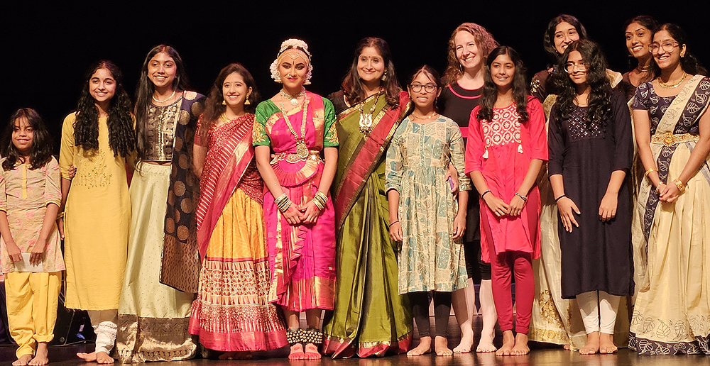 A row of young women and girls, all wearing traditional Indian garments: colorful silken saris or long tunics with leggings. The girl in the center has just finished her debut recital.
