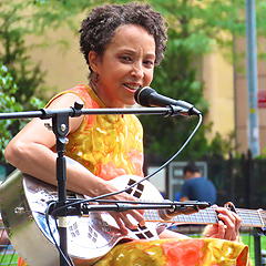 A Black woman on stage, singing and playing a silver resonator guitar. She has short curly hair and is wearing a red print sundress.